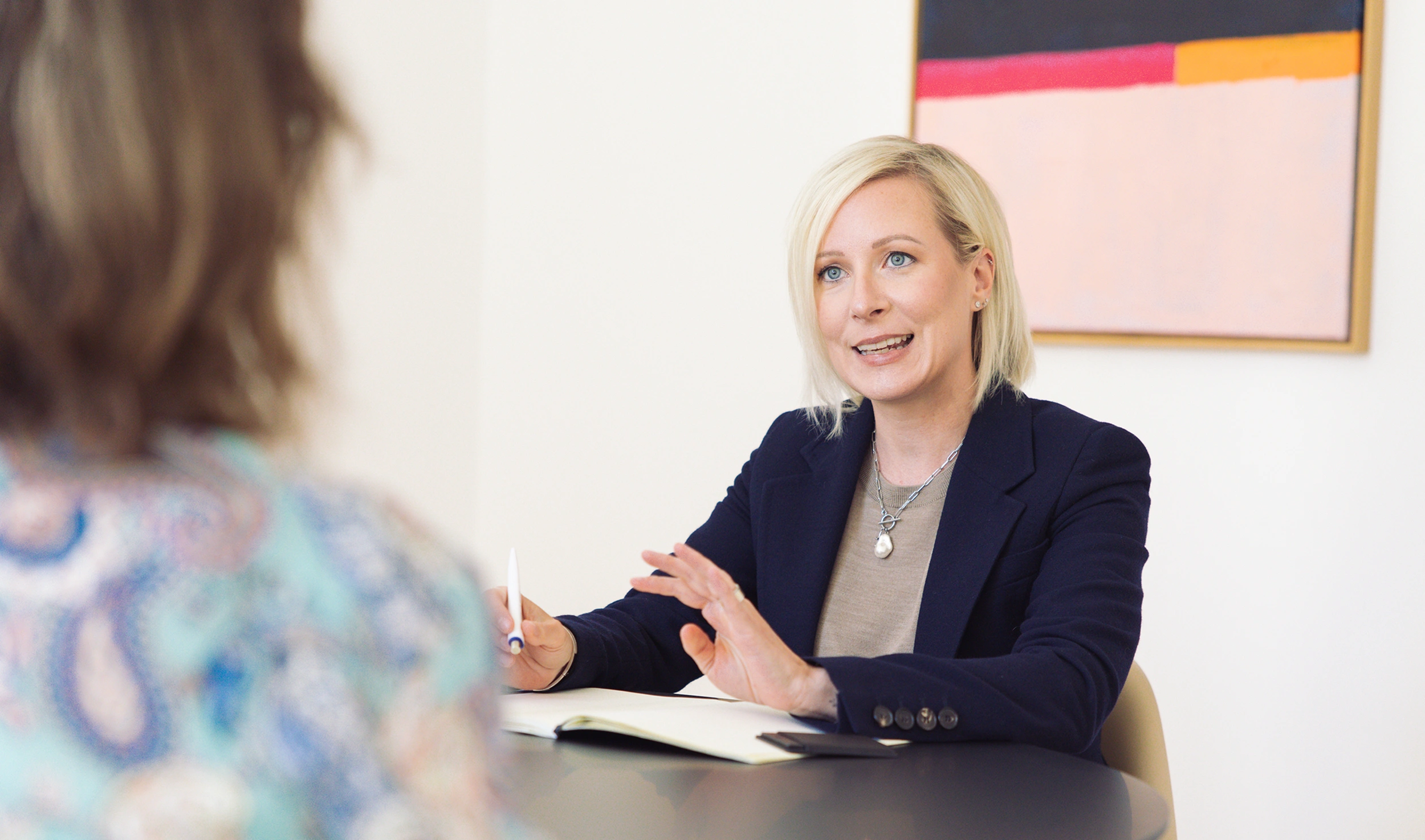 Blonde woman in a navy blazer speaking at a meeting table, gesturing with her hands while another person sits across.