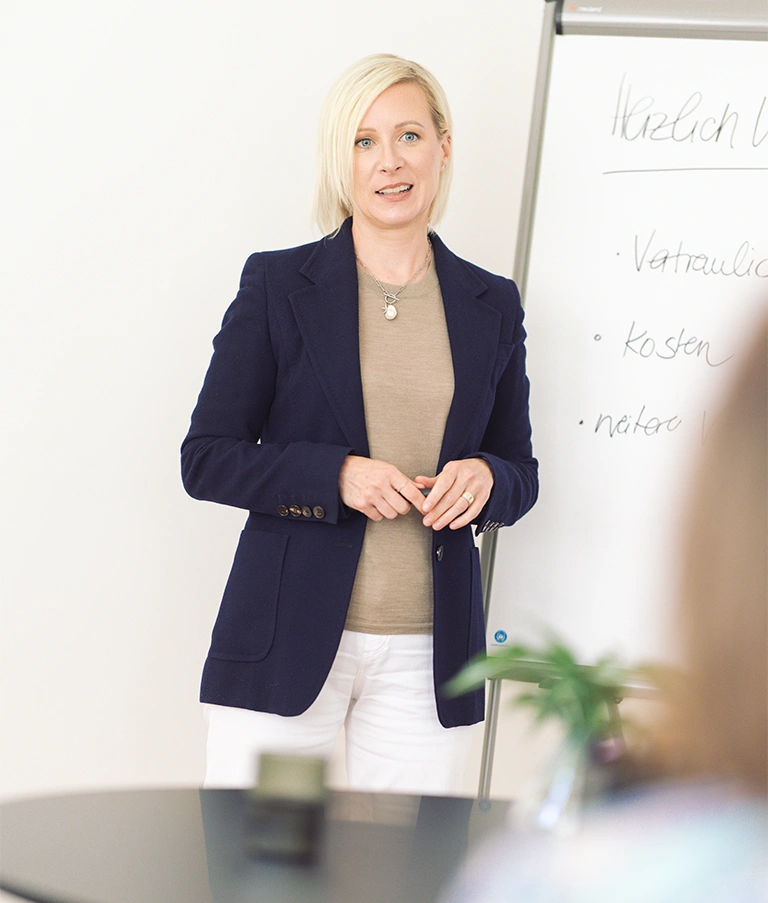 Professional woman in a navy blazer speaks during a workshop, standing by a whiteboard with notes in the background.